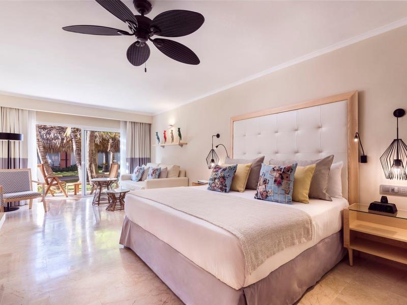 Camera luminosa con letto grande, ventilatore a soffitto e accesso al balcone con area salotto.