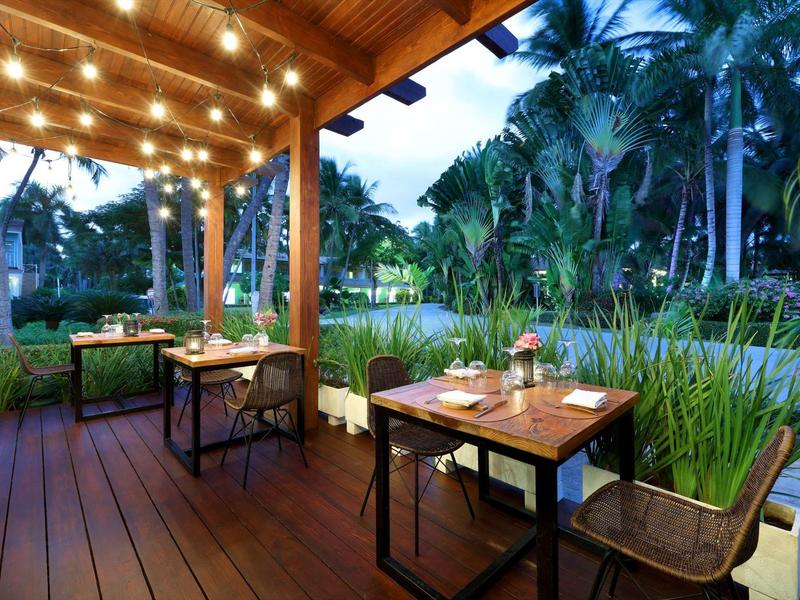 Accogliente area esterna di un ristorante con pavimento in legno, tavoli e luci calde vicino all'acqua e alle palme.