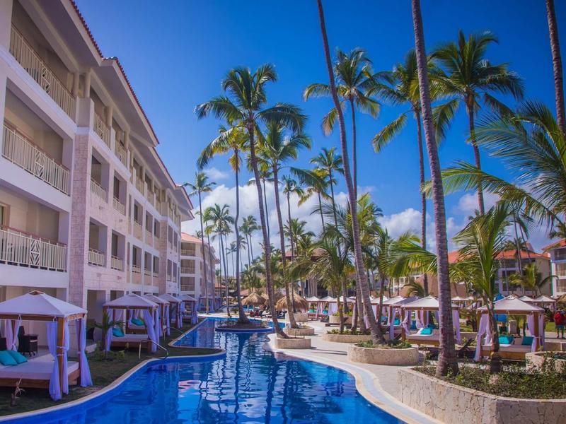 Moderní hotelový komplex s bazénem, palmami a modrou oblohou za slunečného dne.