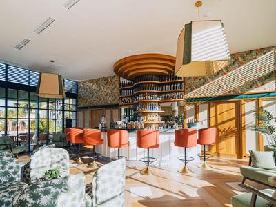 Moderne Hotelbar mit orangen Barhockern, runden Holzdecken-Elementen und großen Fenstern für viel Licht.