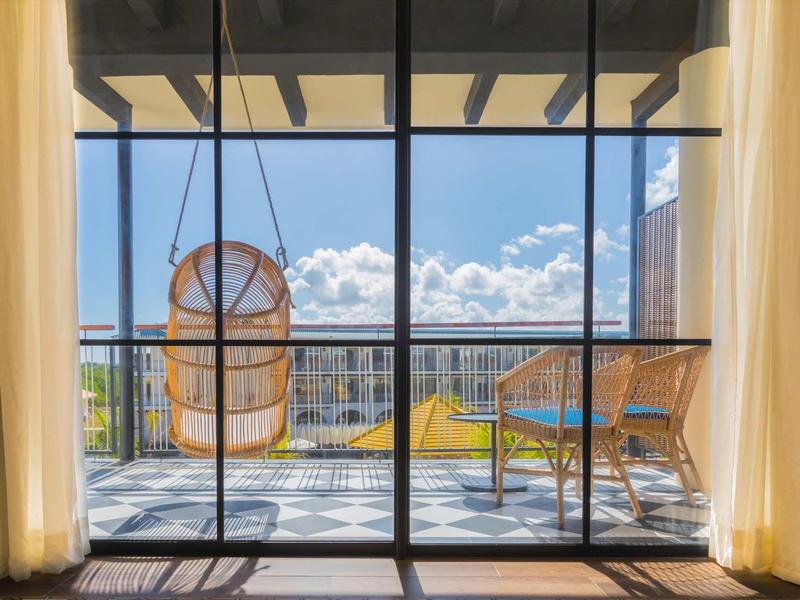Balkon mit Schaukelsessel und Stuhl, Blick auf Stadt und blauen Himmel, helle Vorhänge rahmen die Tür.