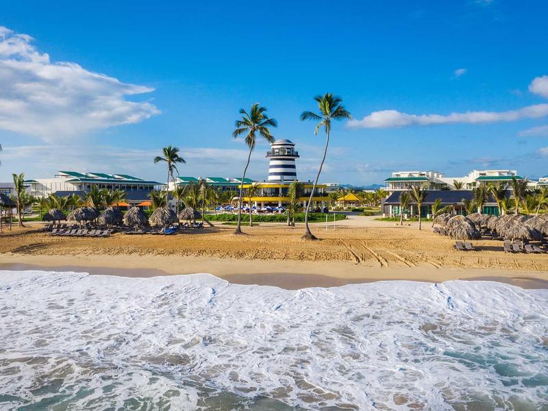 Strand mit goldfarbenem Sand, Palmen, Sonnenschirmen und blauem Himmel mit Wolken.