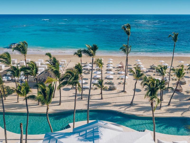 Strand mit Palmen, weißen Sonnenschirmen und blauem Meer im Hintergrund an einem sonnigen Tag.