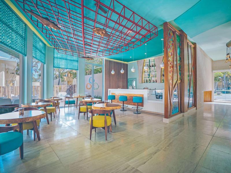 Modernes, helles Restaurant mit großen Fenstern, bunten Stühlen und dekorativem Deckennetz.