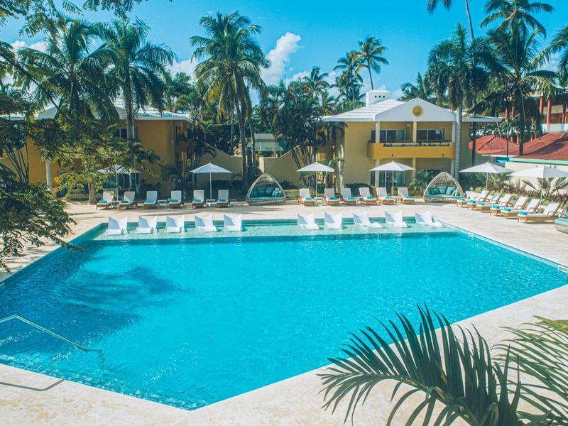 Großer blauer Pool mit weißen Liegestühlen, Palmen und gelben Bungalows bei klarem Himmel.