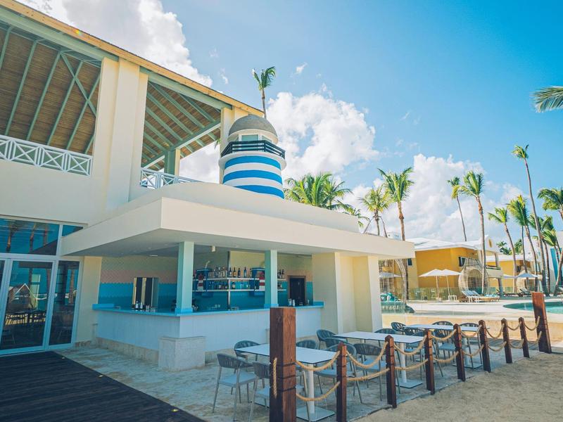 Strandbar mit weiß-blauem Turm, Holzsteg und Palmen unter blauem Himmel am Sandstrand.