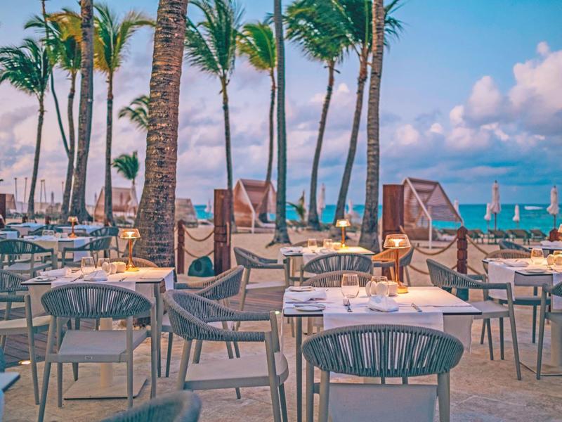 Leerer Strandbereich mit Stühlen, Tischen und Palmen unter blauem Himmel am Meer.