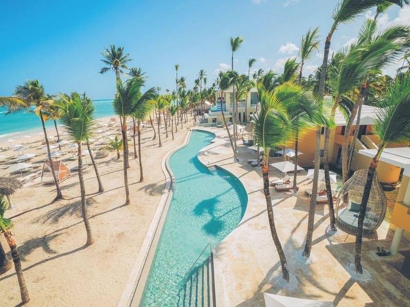 Langer, geschwungener Pool mit Palmen und Sandstrand am türkisfarbenen Meer.