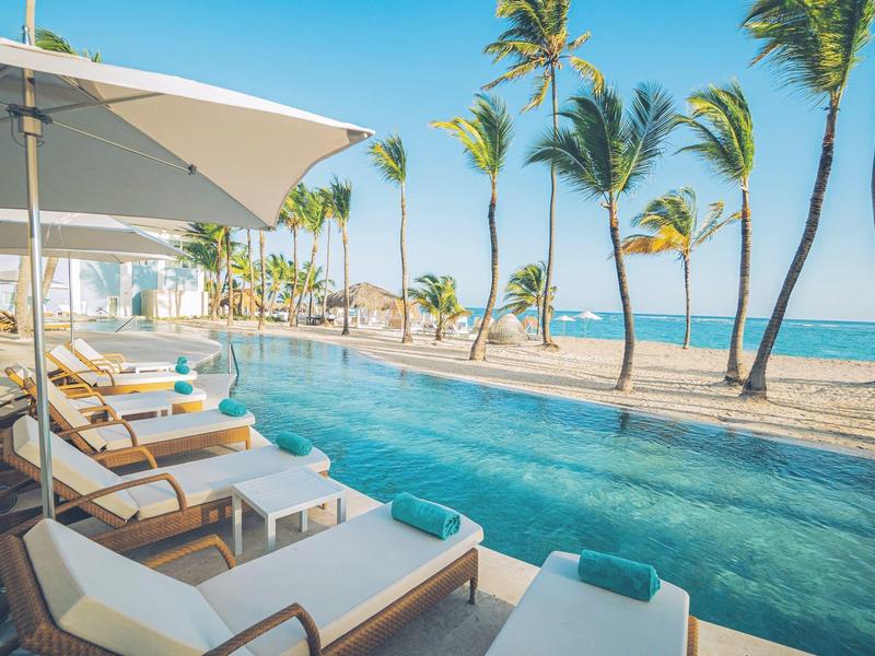 Poolbereich mit Liegen und Sonnenschirmen neben Palmen am Strand unter blauem Himmel.