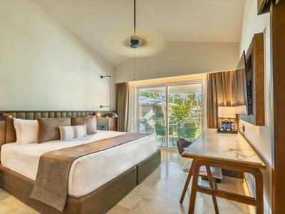 Modern hotel room with large bed, desk, and view of balcony with greenery.