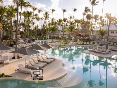 Large pool area with many lounge chairs, surrounded by palm trees and warm sunlight.