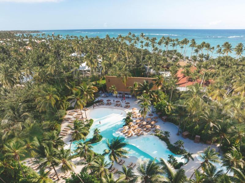 Pohled na tropický hotelový komplex s bazénem, palmami a mořem v pozadí.