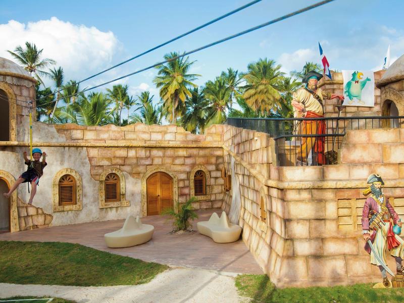 Barevný zábavní park s kamennými zdmi ve tvaru pevnosti a tropickými palmami v pozadí.