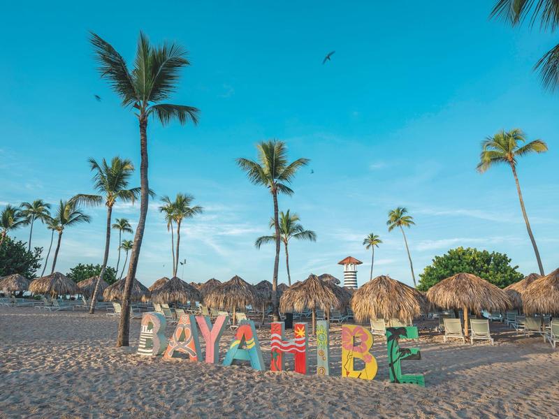 Pláž s barevnými písmeny "BAYAHIBE", palmami a sluncem v pozadí