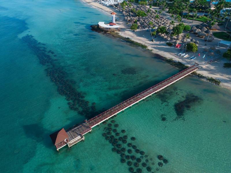 Dlouhý mol vede přes tyrkysovou vodu ke břehu s majákem a vegetací.