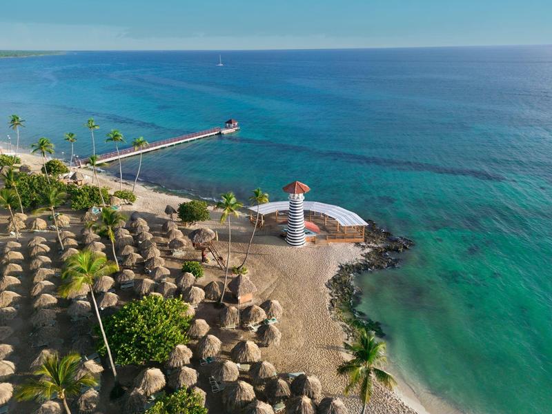 Pláž s mnoha slunečníky, palmami, majákem a molem u čistého modrého moře.