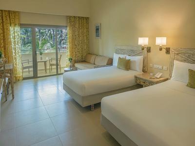 Jasny pokój hotelowy z dwoma łóżkami, kanapą i dostępem do balkonu z widokiem na ogród.