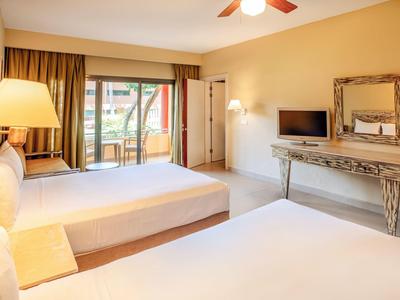 Helles Hotelzimmer mit zwei Doppelbetten, großem Spiegel, TV und Balkon mit Geländer.
