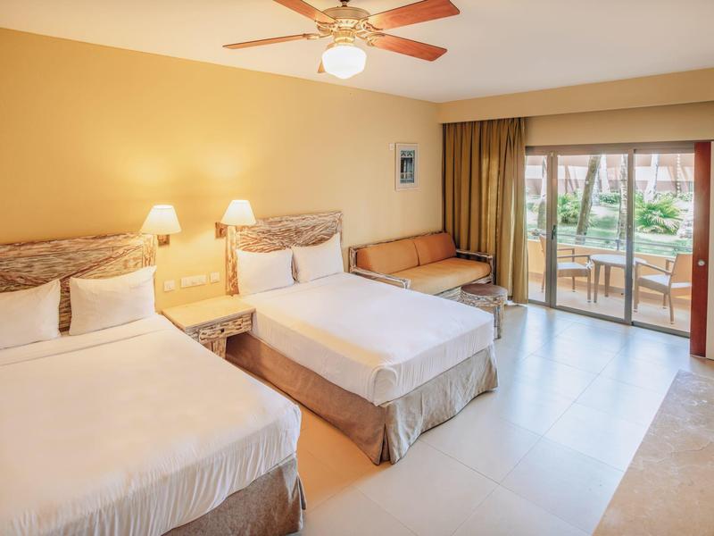 Pokój hotelowy z dwoma łóżkami, lampkami nocnymi, wentylatorem sufitowym i tarasem z widokiem na ogród.