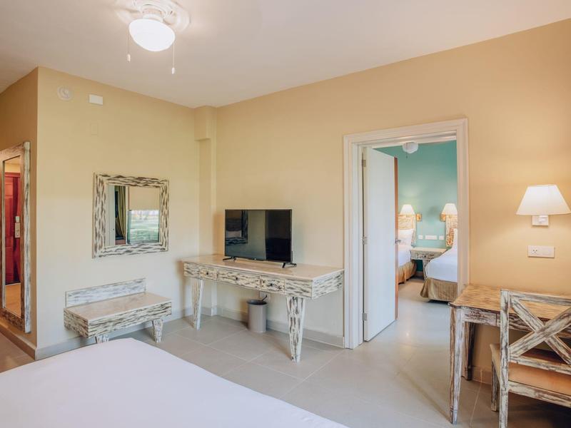 Jasny pokój hotelowy z podwójnym łóżkiem, telewizorem, lustrem, biurkiem i łączonymi drzwiami do sąsiedniego pokoju.