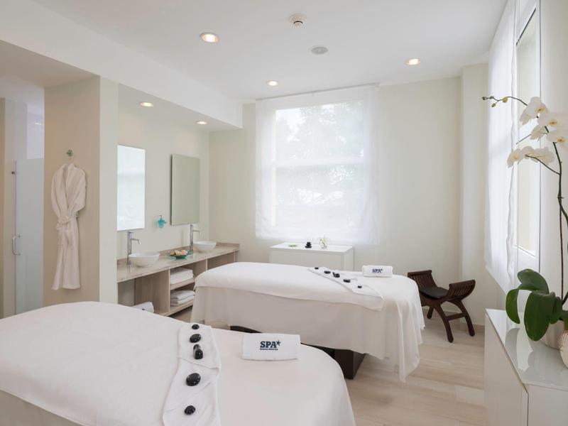 Jasny pokój zabiegowy z dwoma stołami do masażu, białe ściany i delikatne dekoracje.