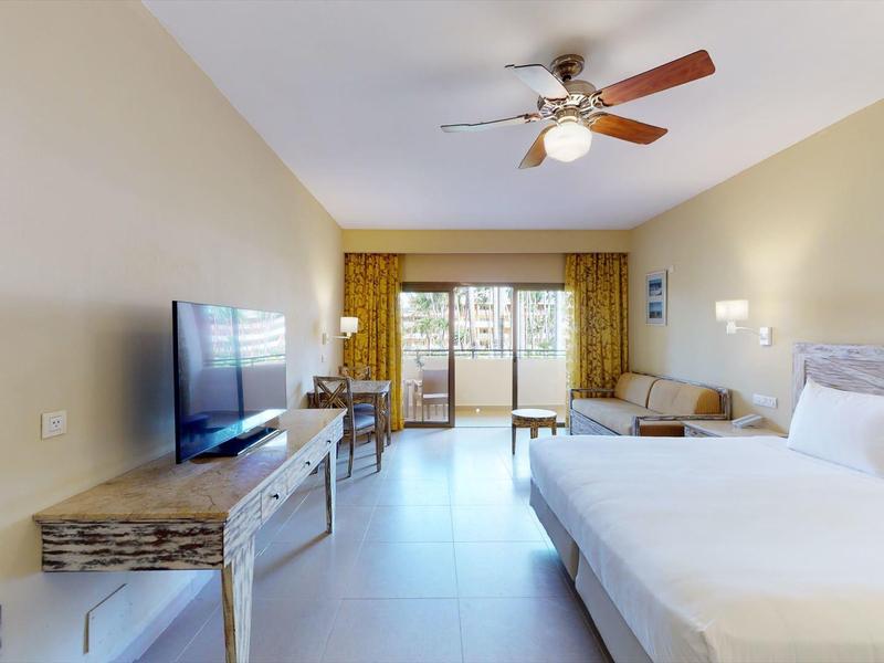 Jasny pokój hotelowy z łóżkiem, telewizorem, wentylatorem sufitowym i balkonem z widokiem na palmy.