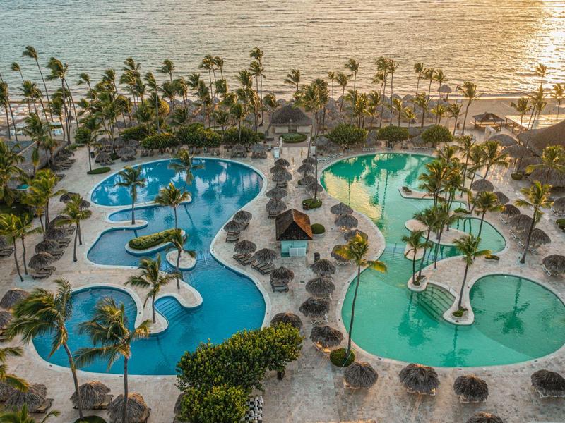 Widok na dwa zakrzywione baseny z palmami i plażą w tle o zachodzie słońca.