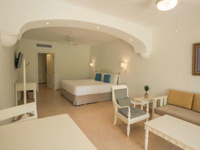 Bright hotel room with double bed, sofa, chairs, and table in neutral colors.