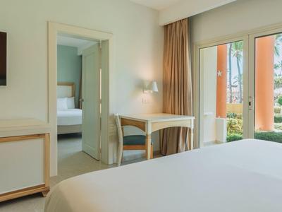 Bright hotel room with double bed, desk, chair, and view of a terrace.