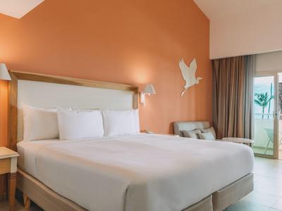 Bright hotel room with large bed, orange wall, and balcony overlooking the sea.