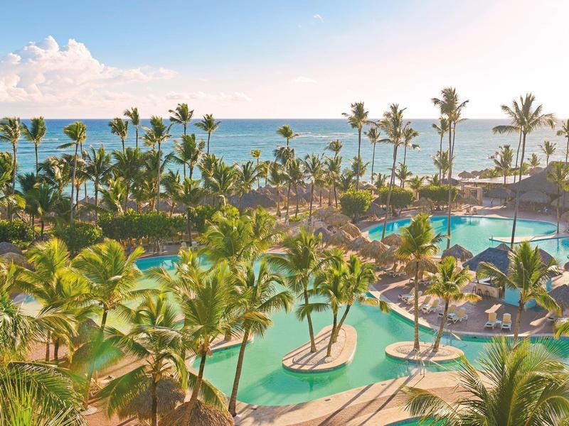 Large tropical hotel pool with palm trees and sea view in sunlight.