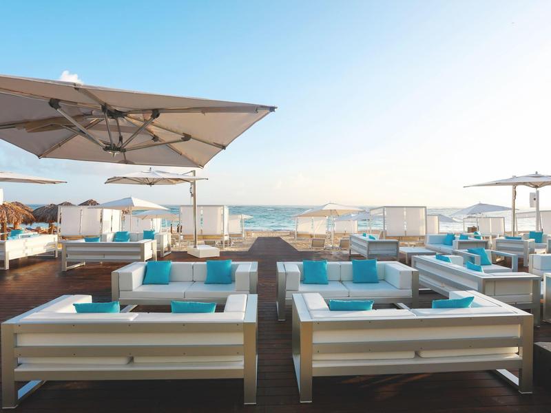 Beach bar with white sofas and large umbrellas, sea view at sunset.