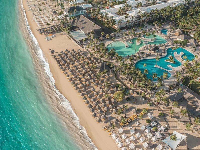 Aerial view of a resort with pool, palm trees, and sandy beach with sun loungers.