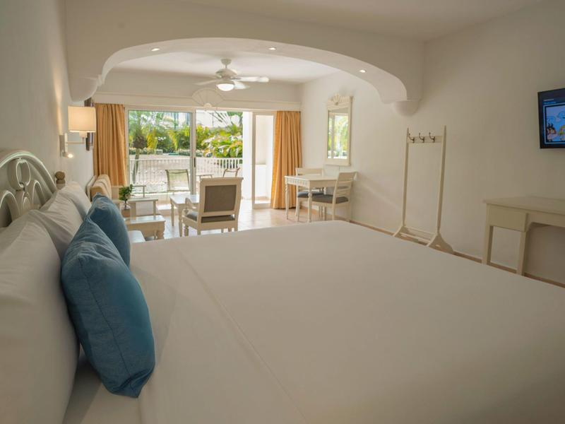 Bright hotel room with large bed, two blue pillows, desk, and balcony overlooking garden.