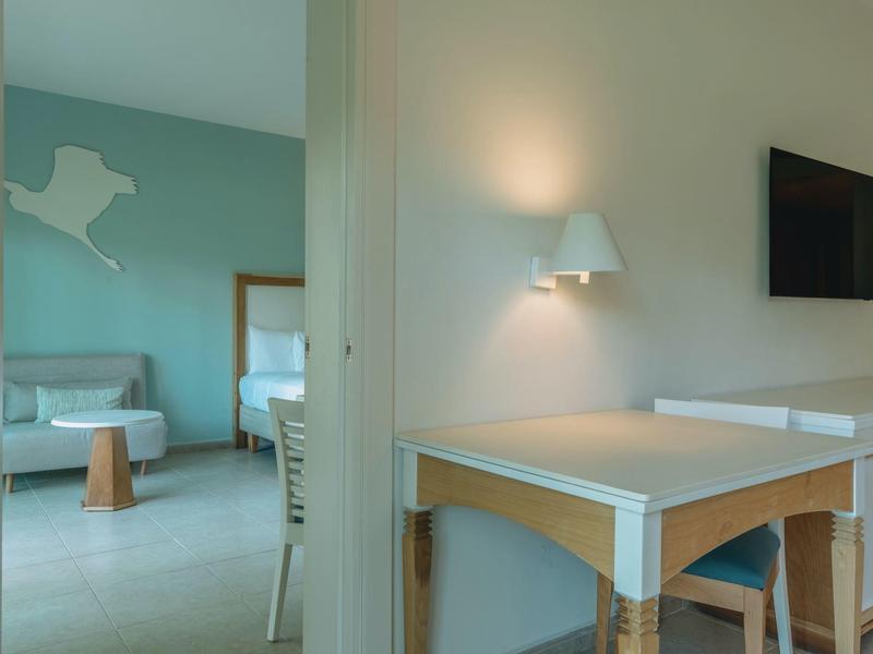 Bright hotel room with desk, chair, and view into bedroom with bed and sofa.