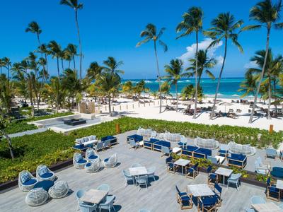 Strandterrasse mit blauen Sesseln, Tischen und grünen Palmen unter klarem blauem Himmel am Meer.