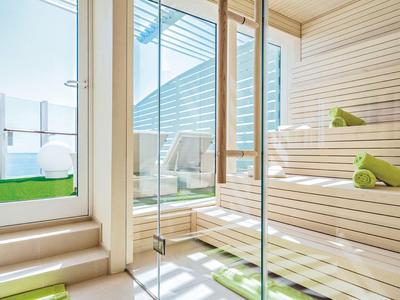 Helle Sauna mit Holzbänken und grünen Handtüchern, Sonnenlicht fällt durch Glasfenster.