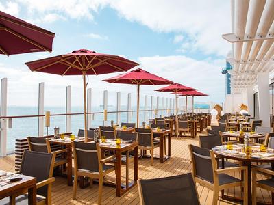 Terrasse mit Tischen und Stühlen, roten Sonnenschirmen und Meerblick bei bewölktem Himmel.