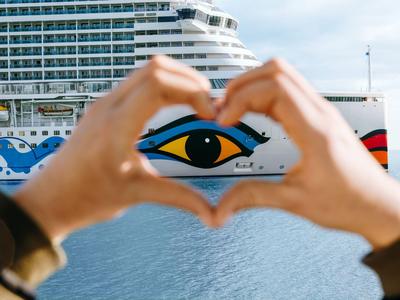 Hände formen ein Herz vor einem großen Kreuzfahrtschiff mit einem bemalten Auge auf dem Rumpf.