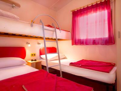 Mały pokój z łóżkiem piętrowym i pojedynczym, czerwona i biała pościel, różowa zasłona.