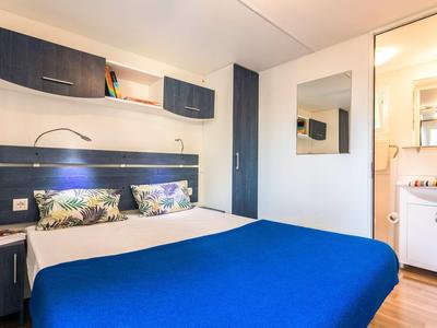 Nowoczesny pokój hotelowy z niebieskim łóżkiem, stolikiem nocnym i dużym lustrem obok łazienki.