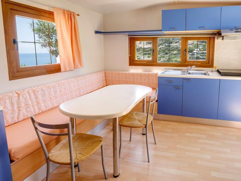 Jasna kuchnia z niebieskimi szafkami, drewnianą podłogą, stołem, krzesłami i oknem z widokiem na morze.