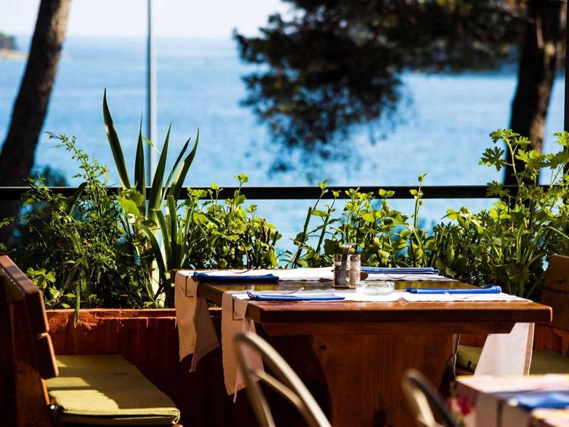 Taras restauracyjny z stołem i krzesłami, otoczony roślinami z widokiem na morze.