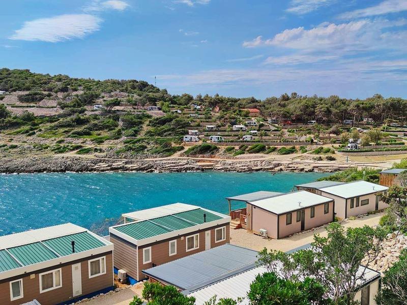 Rząd małych domków letniskowych nad wodą z zielonymi dachami i wzgórzami w tle.