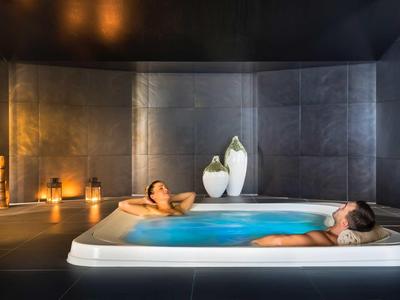 Zwei Personen entspannen in einem modernen Whirlpool, umgeben von dunklen Wänden und warmem Kerzenlicht.