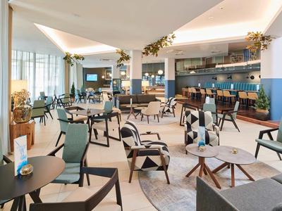 Hell eingerichteter Hotel-Loungebereich mit Sesseln, Tischen und einer Bar im Hintergrund.