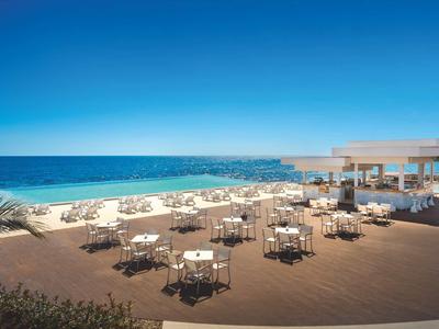 Terrasse mit Tischen und Stühlen am Meer unter klarem blauem Himmel.