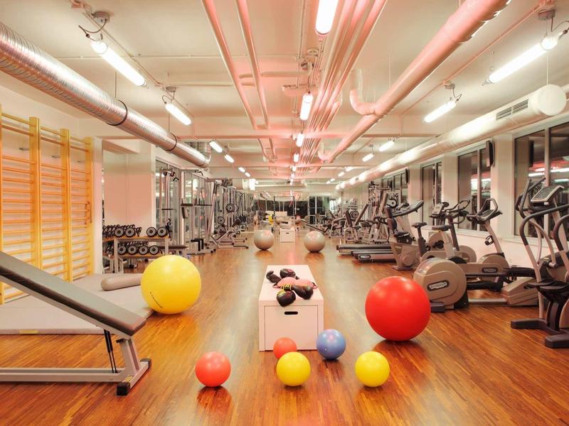 Großer, heller Fitnessraum mit Hanteln, Trainingsgeräten und bunten Gymnastikbällen auf Holzboden.