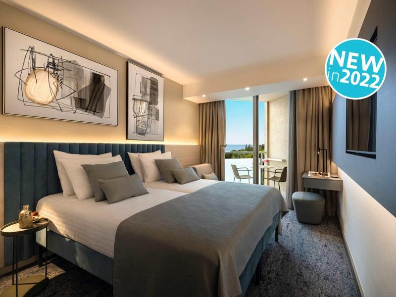 Modernes Schlafzimmer mit großem Bett, grauen Textilien, Kunstbildern und Balkon mit Meeresblick.