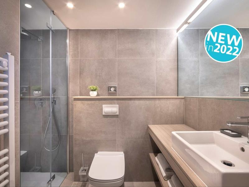 Modernes Badezimmer mit grauen Fliesen, Walk-in-Dusche, weißer Toilette und Waschbecken mit Holzablage.